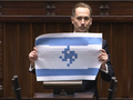 Konrad Berkowicz trzyma plakat z flagą, stojąc przy mównicy w sali obrad