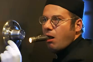 Mężczyzna w okularach trzyma latarkę w ustach, dotyka sejfu białą rękawiczką.