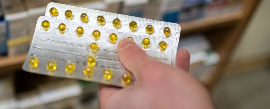 Osoba trzyma opakowanie z żółtymi kapsułkami w aptece. W tle półki z lekami.
