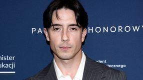 Mężczyzna w przyciemnionym garniturze i białej koszuli, na tle ciemnoniebieskim.