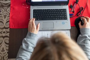 Osoba pracująca na laptopie przy biurku, z notesem i myszką obok. Czerwony obrus pod sprzętem.