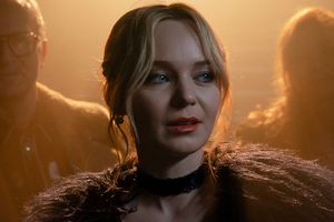 Kobieta z blondu włosami i czerwonymi ustami, w połowie obrócona, w tle rozmyte sylwetki.