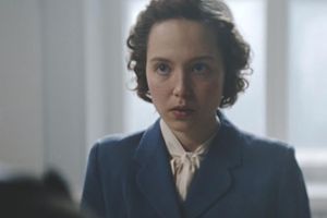 Kobieta w niebieskiej marynarce z zamyśloną miną, w tle jasne okno.