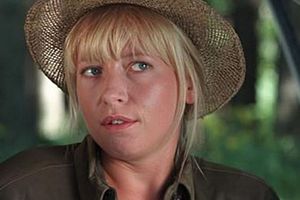 Kobieta z blond włosami w słomkowym kapeluszu, stoi w otoczeniu zieleni, z wyrazem twarzy zastanawiającym.