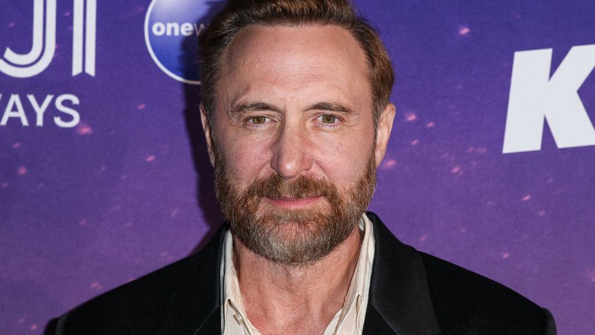 David Guetta z brodą w czarnej marynarce na tle fioletowej ścianki z logo.