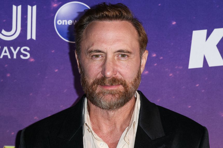 David Guetta z brodą w czarnej marynarce na tle fioletowej ścianki z logo.
