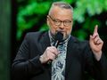 Piotr Bałtroczyk. Mężczyzna z brodą, w okularach, trzyma mikrofon, mówi na tle zielonych liści.