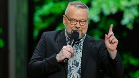 Piotr Bałtroczyk. Mężczyzna z brodą, w okularach, trzyma mikrofon, mówi na tle zielonych liści.