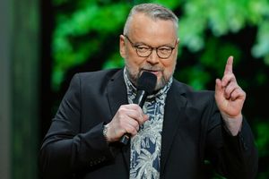 Piotr Bałtroczyk. Mężczyzna z brodą, w okularach, trzyma mikrofon, mówi na tle zielonych liści.