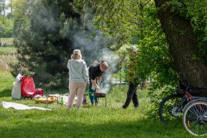 Grupa ludzi grilluje w parku, w otoczeniu zieleni i drzew. Widać dym z grilla.