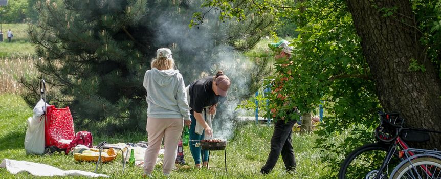 Grupa ludzi grilluje w parku, w otoczeniu zieleni i drzew. Widać dym z grilla.