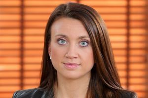 Kobieta z długimi brązowymi włosami, w jasnym topie, patrzy prosto w kamerę. Tło: drewniane żaluzje.