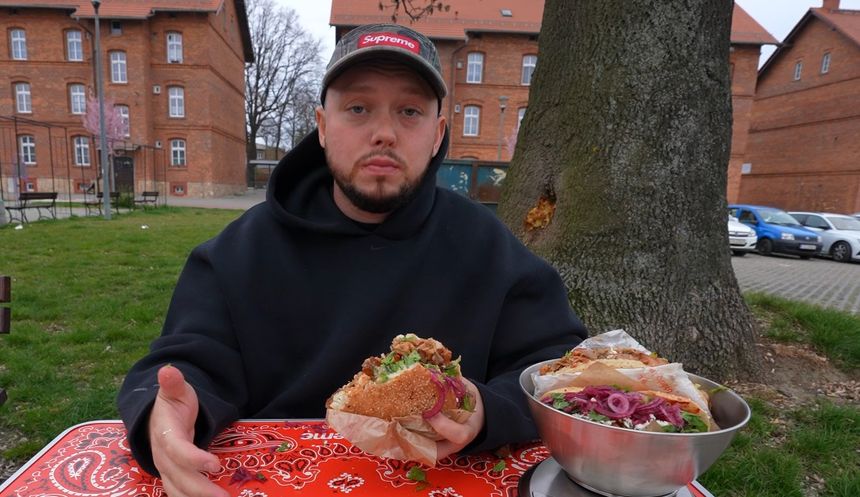 Książulo w czarnej bluzie siedzi przy stole, trzymając kebab.