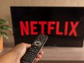 Osoba trzymająca pilot przed telewizorem z logo Netflix na ekranie. W tle roślina doniczkowa.