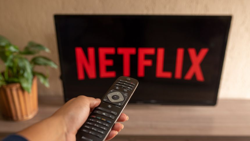 Osoba trzymająca pilot przed telewizorem z logo Netflix na ekranie. W tle roślina doniczkowa.