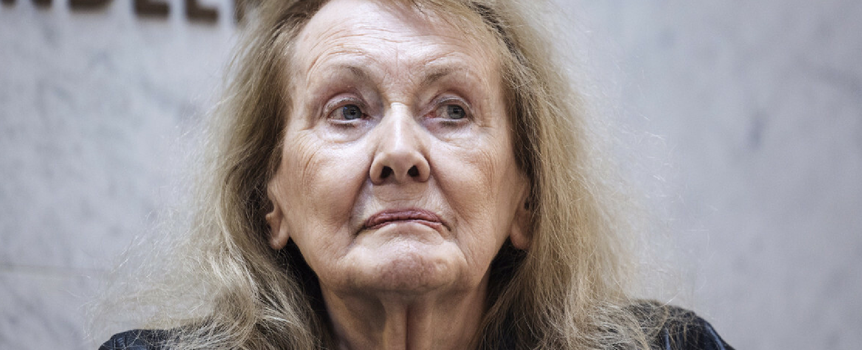 Kobieta z długimi, jasnymi włosami, patrzy w stronę kamery. Tło jednolite, lekko rozmyte.