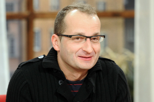 Mężczyzna w okularach, uśmiechnięty, w czarnym swetrze, siedzi w jasnym wnętrzu.