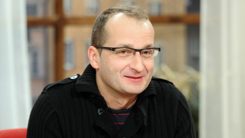 Mężczyzna w okularach, uśmiechnięty, w czarnym swetrze, siedzi w jasnym wnętrzu.