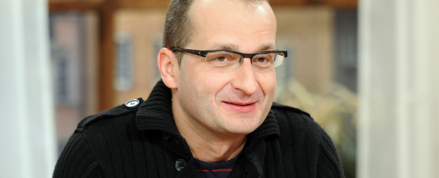 Mężczyzna w okularach, uśmiechnięty, w czarnym swetrze, siedzi w jasnym wnętrzu.