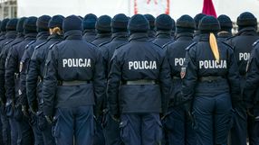 Grupa policjantów w mundurach, stojących na apelu, tyłem do obiektywu, z flagą w tle.