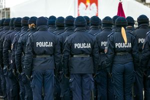 Grupa policjantów w mundurach, stojących na apelu, tyłem do obiektywu, z flagą w tle.