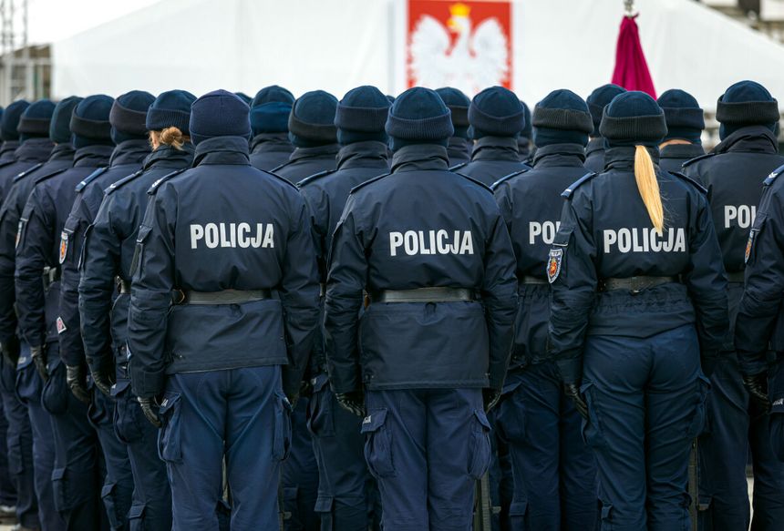Grupa policjantów w mundurach, stojących na apelu, tyłem do obiektywu, z flagą w tle.