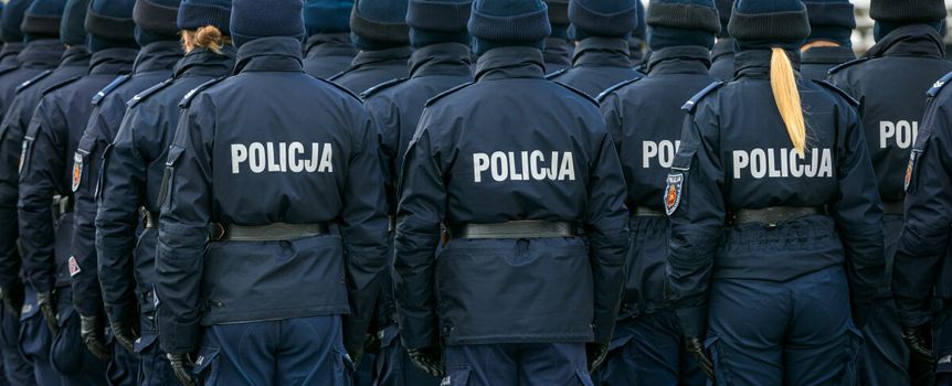 Grupa policjantów w mundurach, stojących na apelu, tyłem do obiektywu, z flagą w tle.