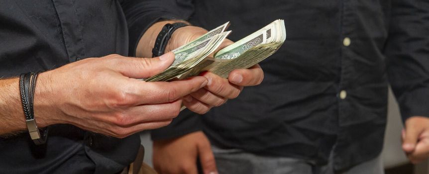 Ręce trzymające banknoty. W tle widoczny mężczyzna w ciemnej koszuli.