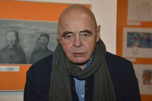 Mężczyzna z łysą głową w szaliku, patrzy w obiektyw, tło z kolorowymi obrazami.