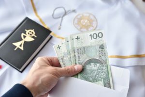 Ręka trzymająca banknoty w kopercie, za nią elementy stroju liturgicznego i książka.