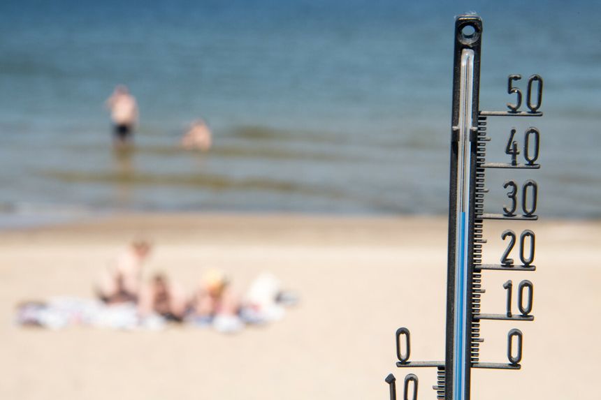Termometr na plaży wskazujący wysoką temperaturę, w tle osoby kąpiące się w wodzie.
