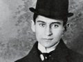 Franz Kafka. Młody mężczyzna w kapeluszu, w formalnym ubraniu, na jednolitym tle. Czarno-biała fotografia.