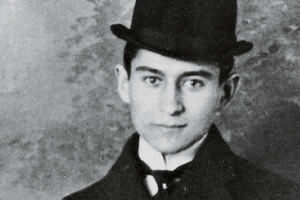 Franz Kafka. Młody mężczyzna w kapeluszu, w formalnym ubraniu, na jednolitym tle. Czarno-biała fotografia.