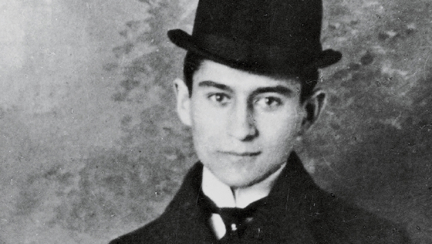 Franz Kafka. Młody mężczyzna w kapeluszu, w formalnym ubraniu, na jednolitym tle. Czarno-biała fotografia.