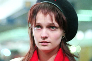 Młoda kobieta z długimi, brązowymi włosami, w beret i czerwoną chustą. Wyraz twarzy poważny.