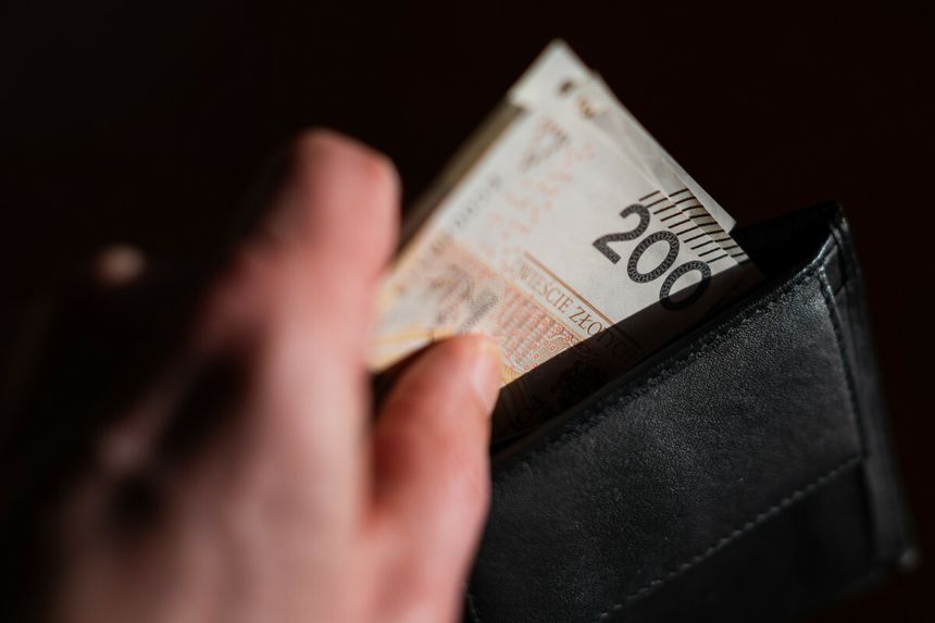 Dłoń wyciąga banknot 200 zł z czarnego portfela na ciemnym tle.