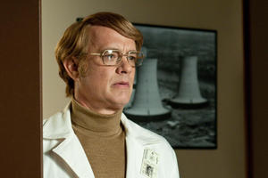 Mężczyzna w białym fartuchu, z okularami, stoi w drzwiach, za nim zdjęcie elektrowni.