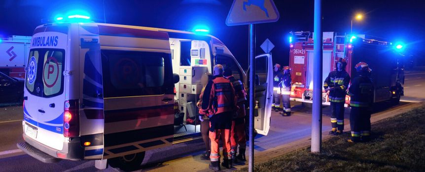 Na zdjęciu karetka i strażacy nocą, w tle niebieskie światła i znak drogowy.