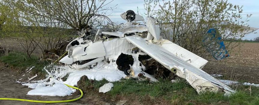 Zniszczony samolot leży w terenie, otoczony roślinnością, widać gaśniczy pianę.