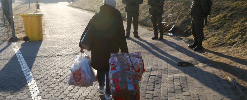Kobieta z zakupami ciągnie wózek, obok stoją żołnierze. Jasna, wczesna pora dnia.