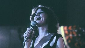 Młoda kobieta śpiewa do mikrofonu, z uśmiechem, w ciemnym stroju. Oklaski w tle.