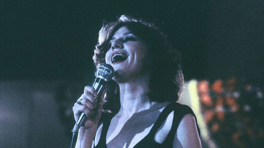 Młoda kobieta śpiewa do mikrofonu, z uśmiechem, w ciemnym stroju. Oklaski w tle.