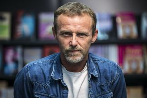 Mężczyzna w niebieskiej kurtce stoi na tle półek z książkami. Ma brodę i poważny wyraz twarzy.