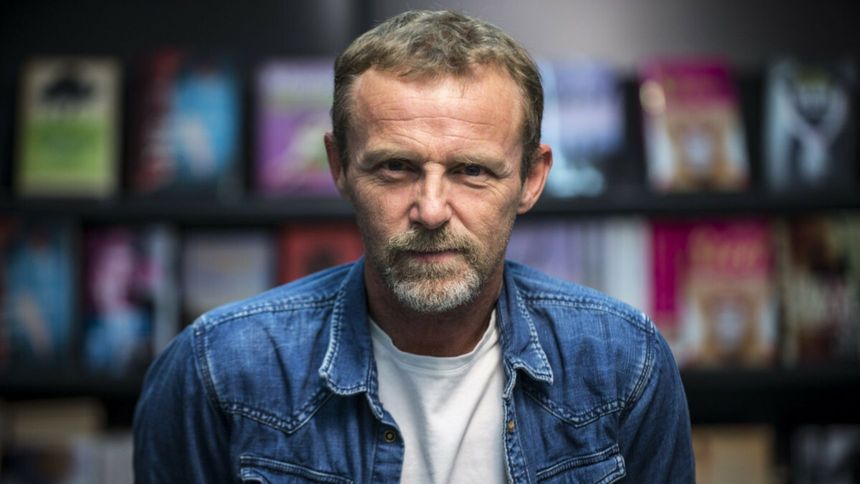 Mężczyzna w niebieskiej kurtce stoi na tle półek z książkami. Ma brodę i poważny wyraz twarzy.