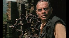 Mężczyzna z przerażonym wyrazem twarzy, trzymający przedmiot w kształcie węży. Zniszczone ruiny w tle.