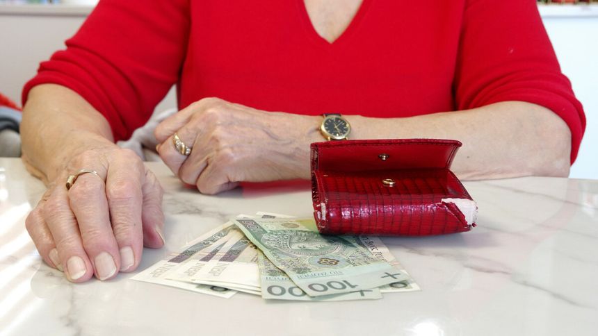Ręce starszej osoby w czerwonym swetrze, z portfelem i banknotami na stole.