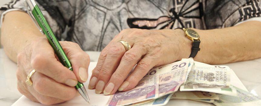 Ręka kobiety piszącej długopisem na kopercie, obok banknoty leżące na stole.