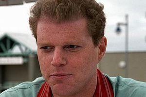 Mężczyzna z jasnymi włosami w niebieskiej koszuli siedzi przy stole na zewnątrz.