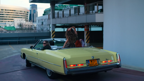 Dwie osoby w żółtym kabriolecie na parkingu, w tle nowoczesne budynki.