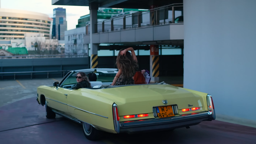 Dwie osoby w żółtym kabriolecie na parkingu, w tle nowoczesne budynki.